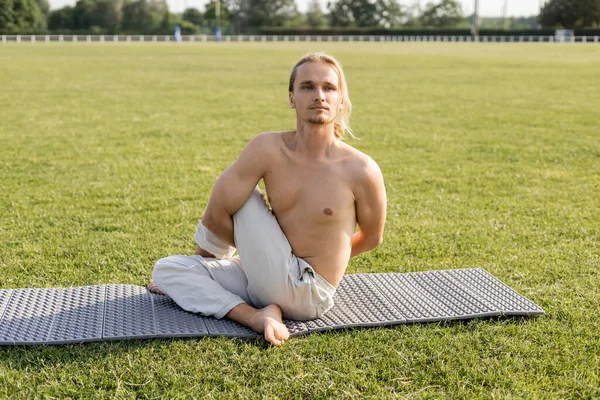 Мужчина без рубашки в хлопковых штанах сидит в мудрой позе и смотрит в сторону, практикуя йогу на зеленой лужайке под открытым небом — стоковое фото