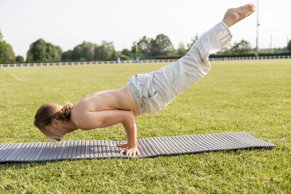 Вид сбоку молодого человека без рубашки в льняных штанах медитирующего в позе павлина на коврике для йоги на зеленой лужайке — стоковое фото