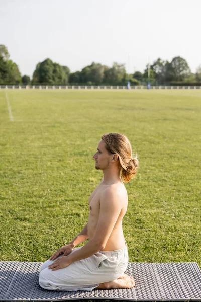 Vista lateral del hombre descalzo sin camisa sentado en la pose del rayo mientras medita en el campo verde en la esterilla de yoga - foto de stock