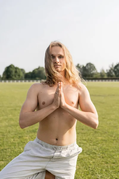 Long haired and shirtless man in linen pants meditating in tree pose with praying hands on green lawn outdoors — Stock Photo