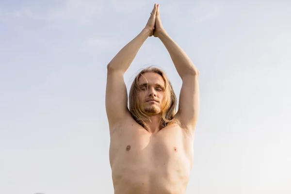 Visão de baixo ângulo do homem sem camisa com cabelos longos e olhos fechados meditando com mãos de oração levantadas contra o céu azul — Fotografia de Stock