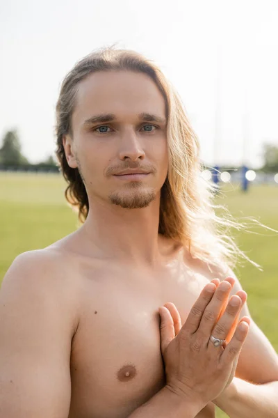 Positive shirtless man with long hair meditating with anjali mudra gesture and smiling at camera outdoors — Stock Photo