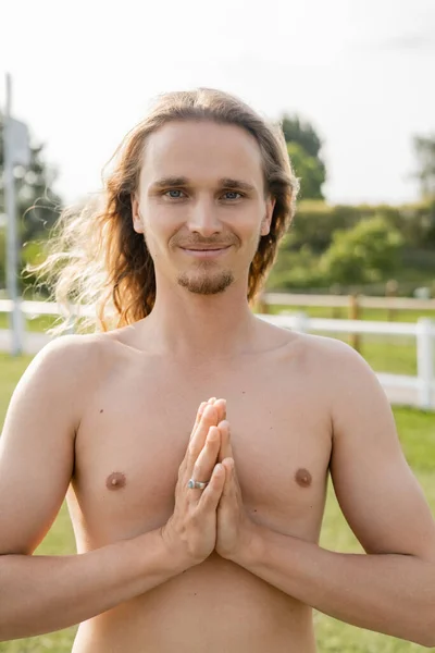 Homme torse nu avec les cheveux longs montrant geste anjali mudra et souriant à la caméra à l'extérieur — Photo de stock