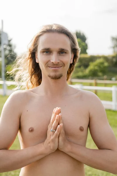 Hombre sin camisa lleno de alegría con el pelo largo mostrando gesto de anjali mudra y sonriendo a la cámara al aire libre — Stock Photo