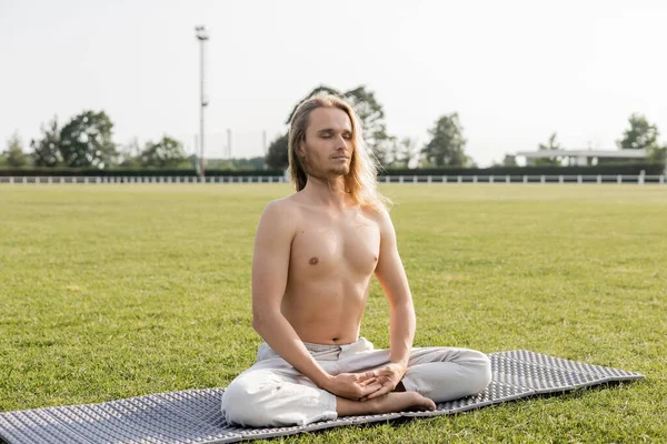 Hombre sin camisa y de pelo largo meditando en pose fácil con los ojos cerrados en la estera del yoga en el estadio al aire libre herboso - foto de stock
