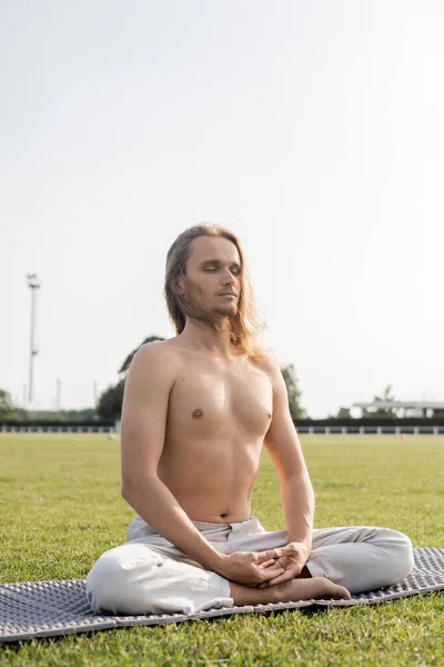 Hombre sin camisa y deportivo con los ojos cerrados meditando en pose de yoga fácil sobre césped verde - foto de stock