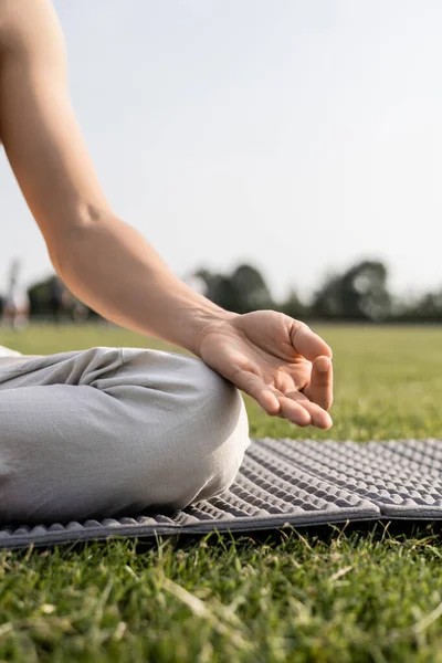 Ausgeschnittene Ansicht eines jungen Mannes, der meditiert und Gyan Mudra Geste zeigt, während er auf einer Yogamatte auf grünem Rasen sitzt — Stockfoto