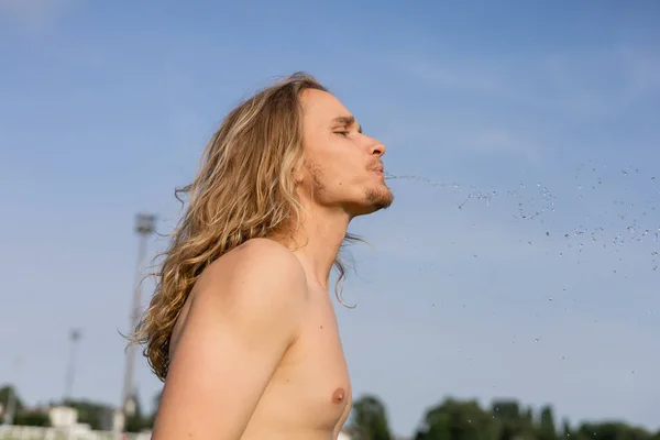 Vista lateral do homem de cabelos longos sem camisa cuspir água doce enquanto faz exercício pranayama ao ar livre — Fotografia de Stock