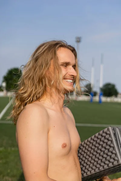 Hombre sin camisa lleno de alegría con pelo largo sosteniendo estera de yoga y sonriendo al aire libre - foto de stock