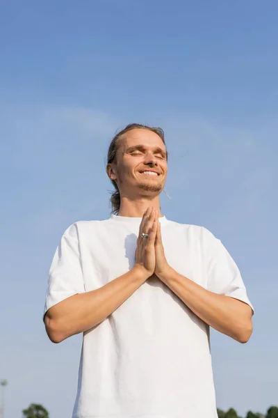Vista de ángulo bajo del hombre alegre en camiseta blanca meditando con los ojos cerrados y las manos rezando bajo el cielo despejado — Stock Photo