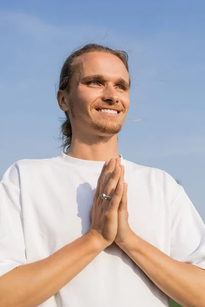 Happy young man in white t-shirt looking away and meditating with praying hands under blue sky — Stock Photo