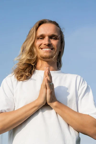 Niedrigwinkel-Ansicht eines lächelnden langhaarigen Yoga-Mannes, der mit betenden Händen meditiert und vor blauem Himmel in die Kamera blickt — Stockfoto