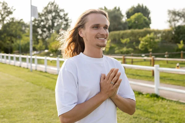 Joyful long haired man in white t-shirt looking away while meditating with anjali mudra outdoors — Stock Photo