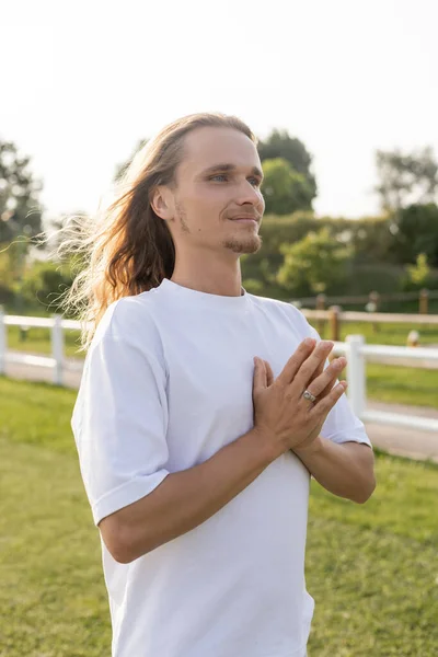 Homem calmo e despreocupado com cabelos longos olhando para longe enquanto meditava com mudra anjali ao ar livre — Fotografia de Stock