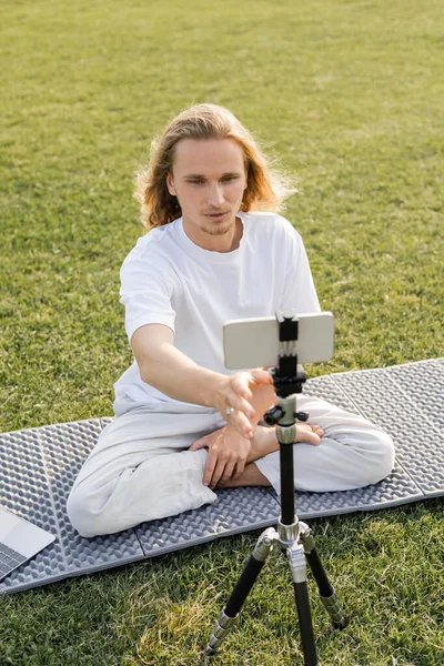 Молодой Vlogger yoga регулируя штатив с мобильным телефоном пока сидя в легкой позе на поле травы — стоковое фото