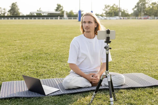 Long haired yoga teacher looking away while sitting near laptop and tripod with mobile phone on field of outdoor stadium — Stock Photo