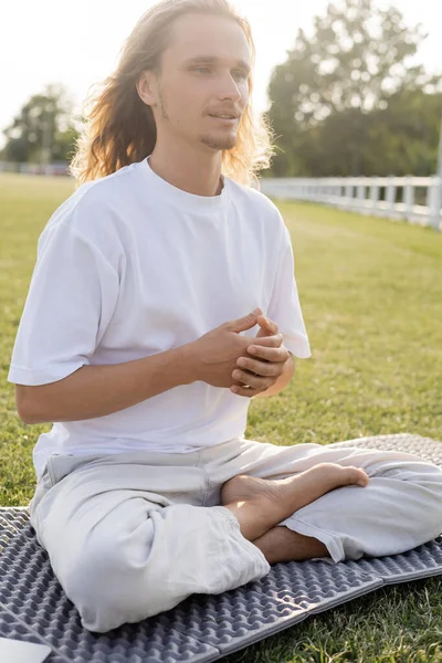 Полная длина босиком мужчина в белой футболке и хлопковых штанах, сидящих в легкой позе и медитирующих на травянистом поле — стоковое фото