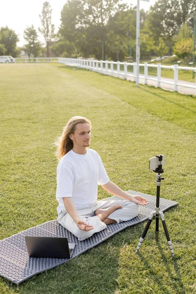 Insegnante di yoga mostrando posa facile e gesti mudra gyan mentre seduto su tappetino yoga vicino smartphone su treppiede all'aperto — Foto stock