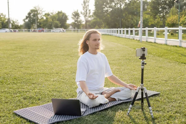 Yoga vlogger méditant dans la pose facile près du téléphone mobile sur le trépied sur la pelouse verte du stade extérieur — Photo de stock