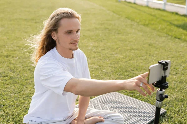 Junger langhaariger Mann benutzt Handy, während er auf Yogamatte auf grünem Rasen sitzt — Stockfoto