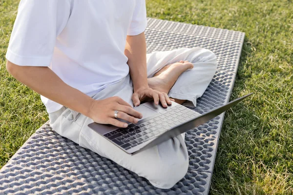 Vue recadrée de l'homme en utilisant un ordinateur portable tout en étant assis en pose facile sur tapis de yoga sur herbe verte — Photo de stock