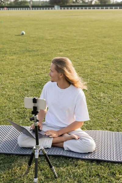 Entrenador de yoga sentado con el ordenador portátil cerca del teléfono celular en el trípode y mirando hacia otro lado en el estadio cubierto de hierba - foto de stock