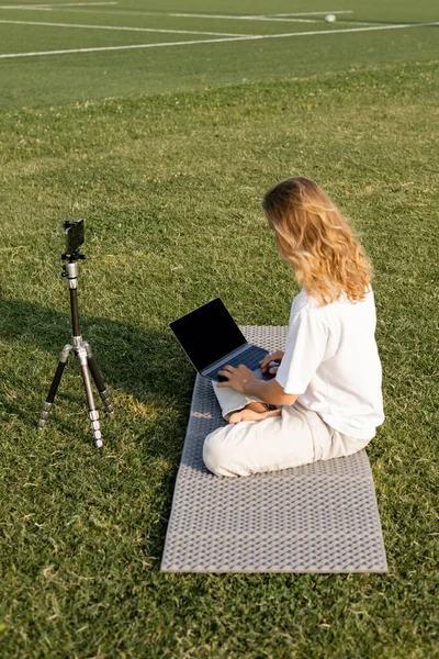 Profesor de yoga de pelo largo usando el ordenador portátil mientras está sentado cerca del trípode con el teléfono celular en el estadio herboso - foto de stock