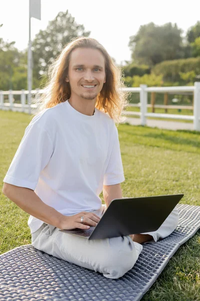 Hombre de pelo largo feliz sentado en pose fácil con el ordenador portátil y sonriendo a la cámara durante la clase de yoga al aire libre - foto de stock
