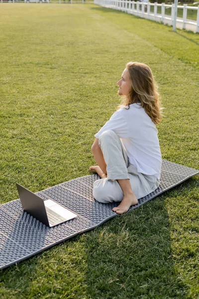 Long haired man sitting in sage pose near laptop and looking away on yoga mat on green field — Stock Photo
