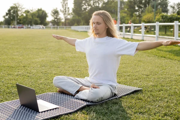 Joven sentado en pose fácil con las manos extendidas durante la lección de yoga en línea en el portátil al aire libre - foto de stock