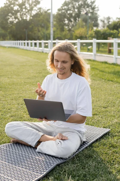 Ein lächelnder Mann spricht während einer Yoga-Stunde am Laptop, während er in lockerer Pose auf dem Außenplatz des Stadions sitzt — Stockfoto