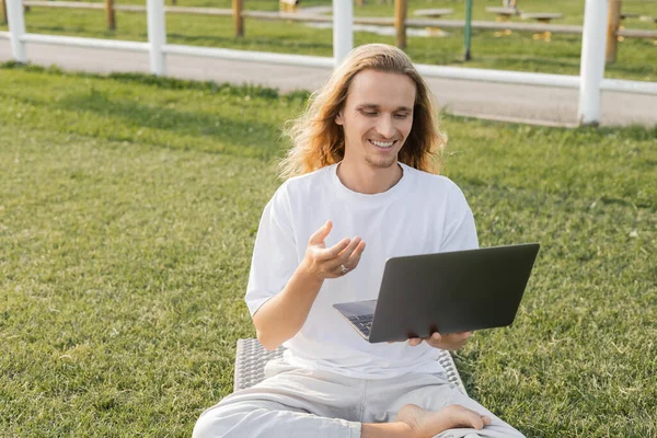 Sonriente hombre de yoga hablando cerca del ordenador portátil durante la lección en línea sobre hierba verde al aire libre - foto de stock