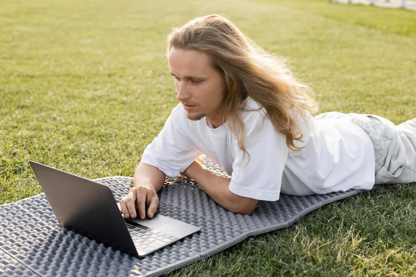 Young long haired man looking at laptop during yoga lesson on green field outdoors — Stock Photo