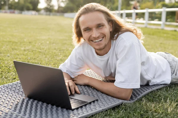 Jeune et insouciant homme regardant la caméra tout en étant couché près d'un ordinateur portable sur tapis de yoga — Photo de stock