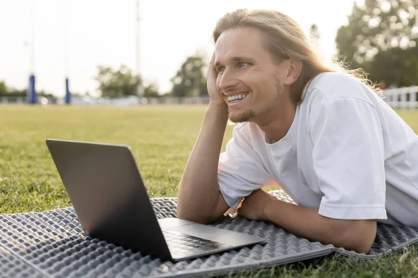 Jovem homem de ioga alegre olhando para longe, enquanto deitado perto de laptop no estádio gramado ao ar livre — Fotografia de Stock