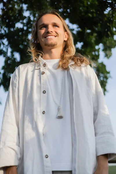 Vista basso angolo di spensierato uomo yoga dai capelli lunghi in abiti di lino sorridente all'aperto — Foto stock