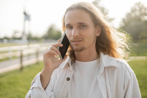 Elegante hombre de yoga de pelo largo mirando a la cámara durante la conversación en el teléfono móvil al aire libre - foto de stock