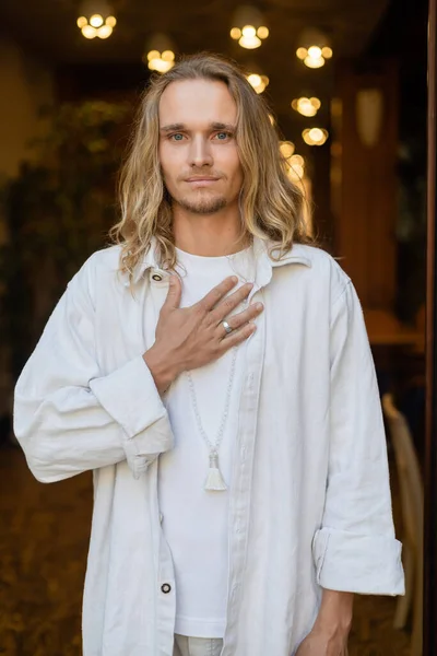 Hellhaariger Yoga-Mann in weißen Leinenkleidern, der die Brust berührt und in die Kamera in der Nähe von Stadtlichtern auf verschwommenem Hintergrund blickt — Stockfoto