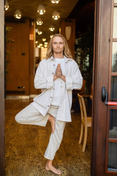Full length of barefoot man in white linen clothes standing in tree pose with praying hands in house with blurred lights — Stock Photo