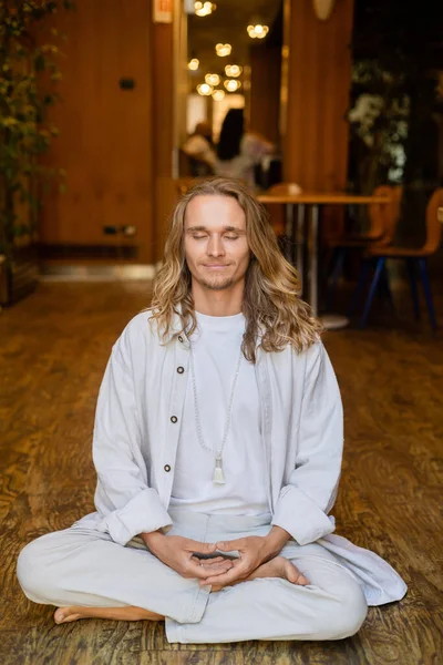 Unbekümmerter Mann mit geschlossenen Augen und langen blonden Haaren meditiert in lockerer Pose im Haus — Stockfoto