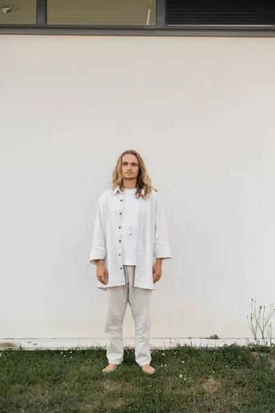 Pleine longueur de pieds nus et homme aux cheveux blonds dans des vêtements en lin debout près du mur blanc à l'extérieur — Photo de stock