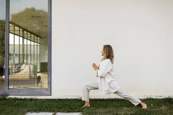 Side view of long haired man in linen clothes meditating in warrior pose with praying hands near white wall of modern cottage — Stock Photo