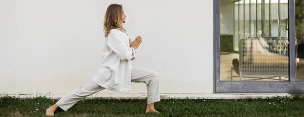 Vista lateral do homem elegante em roupas brancas praticando ioga em pose guerreira com gesto mudra anjali perto da casa moderna, banner — Fotografia de Stock