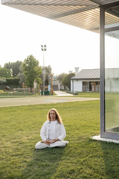 Hipster hombre en ropa blanca sentado en pose de loto durante la meditación sobre hierba verde cerca de la casa de campo - foto de stock