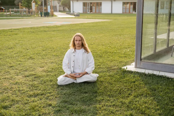 Junger und stilvoller Mann in Leinenkleidung sitzt auf dem Rasen in der Nähe von Hütten und meditiert in Lotus-Pose — Stockfoto