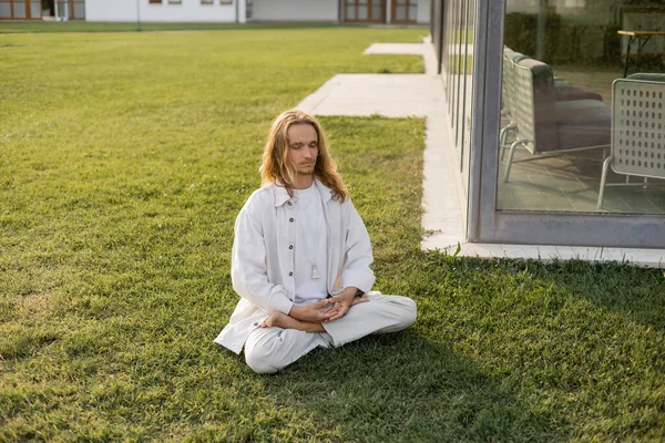 Стильный длинноволосый мужчина медитирует в позе лотоса, сидя на газоне рядом со зданием — стоковое фото