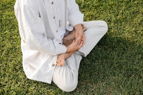 Частичный вид босоногого человека в белой льняной одежде медитирующего в позе лотоса на зеленом газоне — стоковое фото