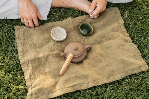 Top view of linen rug with palo santo stick and ceramic teapot with cups near cropped man sitting on green grass — Stock Photo