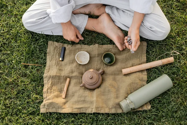Top view of cropped barefoot man in white clothes holding mala beads near linen rug with ceramic cups and teapot on green lawn — Stock Photo