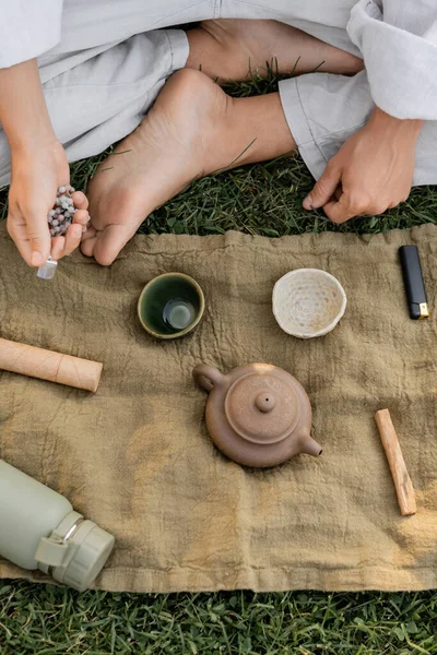Частичный вид босоногого человека с бусинками, сидящего возле льняного ковра с глиняными чашками и чайником на лужайке на открытом воздухе — стоковое фото